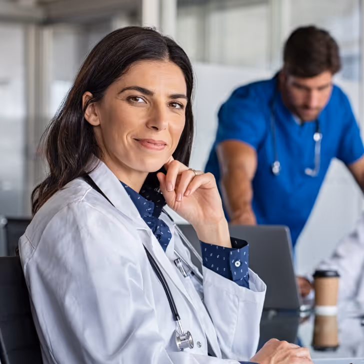 Female doctor in a white coat with a stethoscope, smiling and seated with a male medical professional working on a laptop in the background.