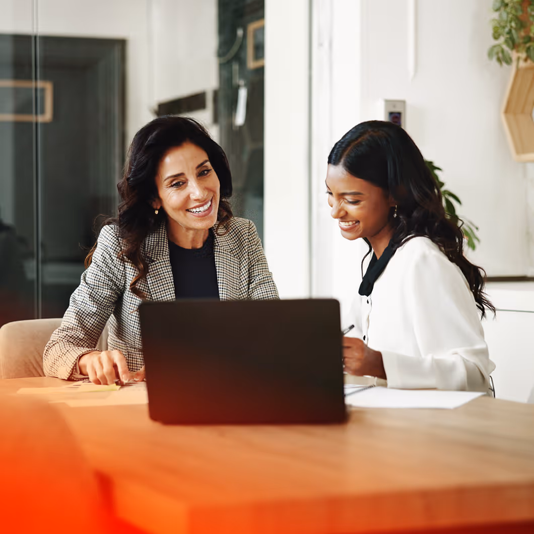 Two women sitting at a table, smiling and looking at a laptop together in an office setting.