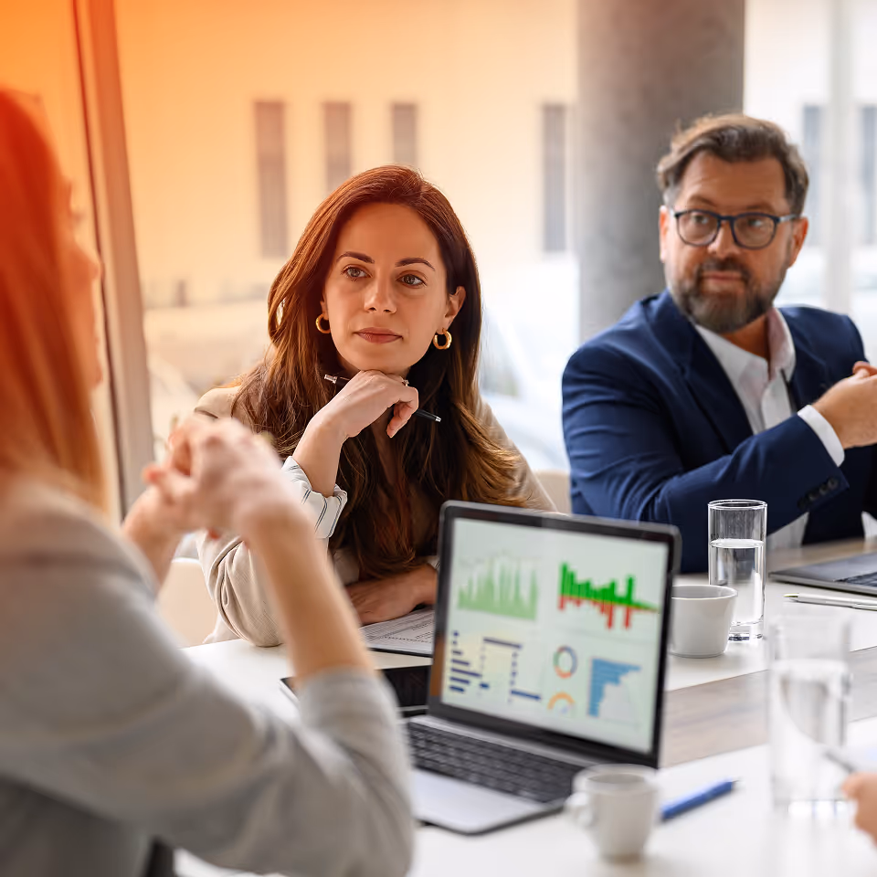 Three professionals in a meeting room with a laptop showing financial charts on the table.