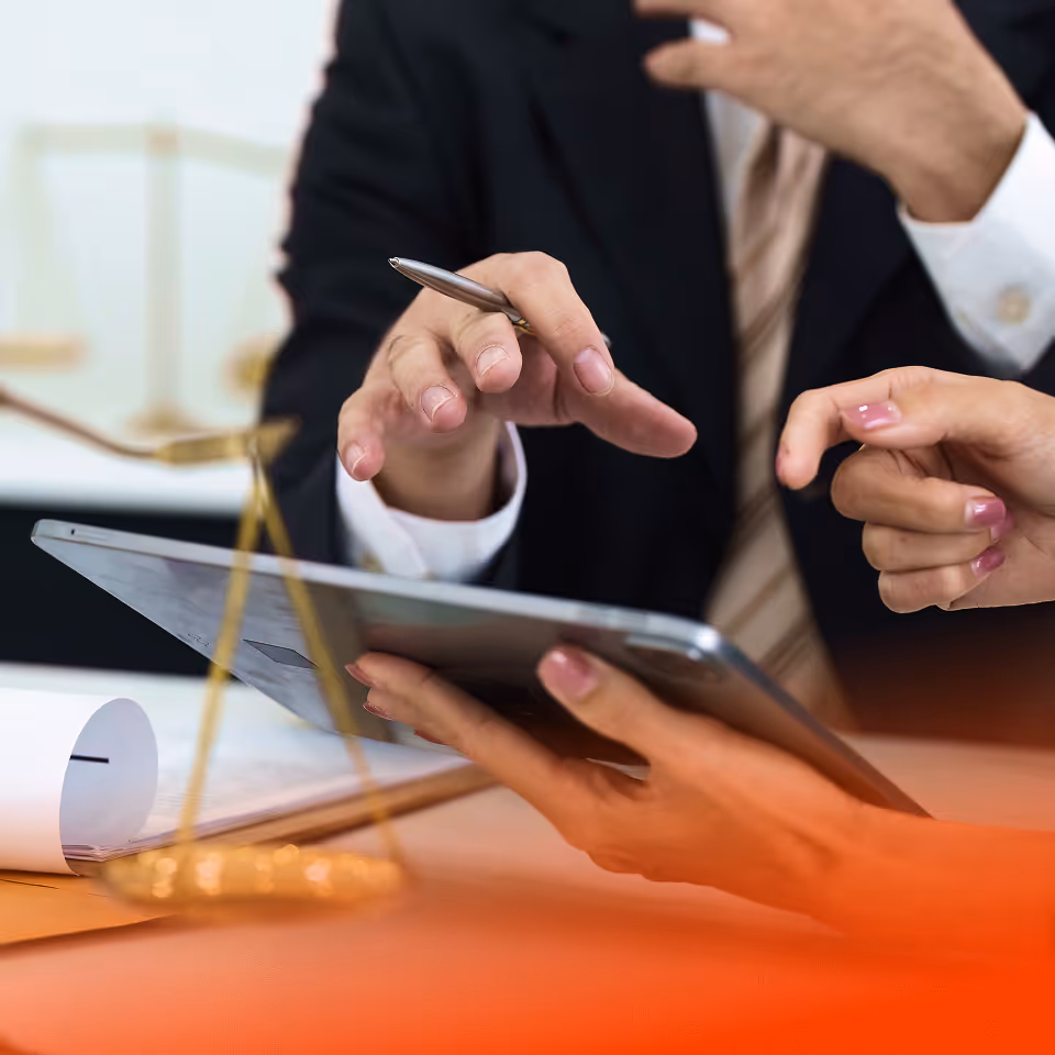 Two people discussing documents while one holds a tablet and the other points with a pen near a golden scale of justice.