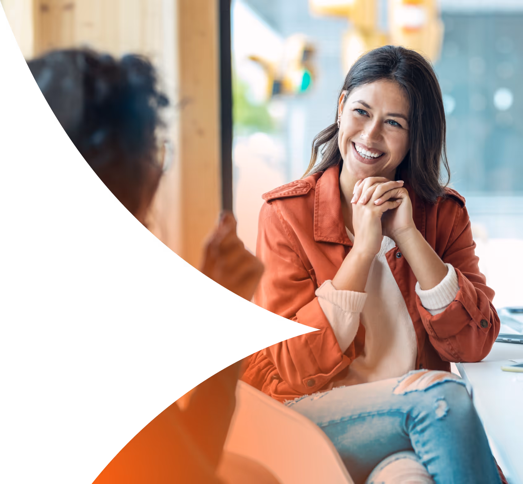 Smiling woman in a red jacket and ripped jeans engaging in conversation with another person in a bright indoor setting.