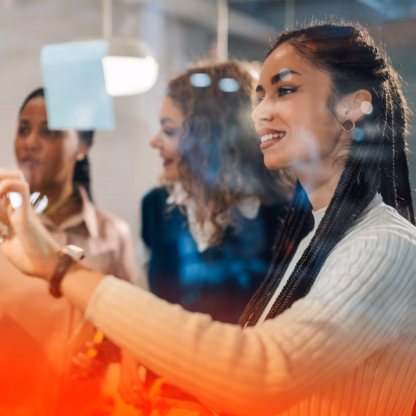 Three women collaborating and smiling while placing sticky notes on a glass wall in an office.