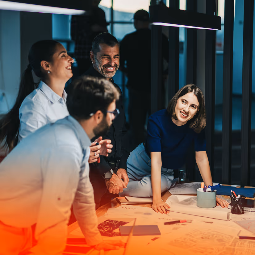 Four colleagues engaged in a lively discussion around a table covered with blueprints and documents in a modern office.