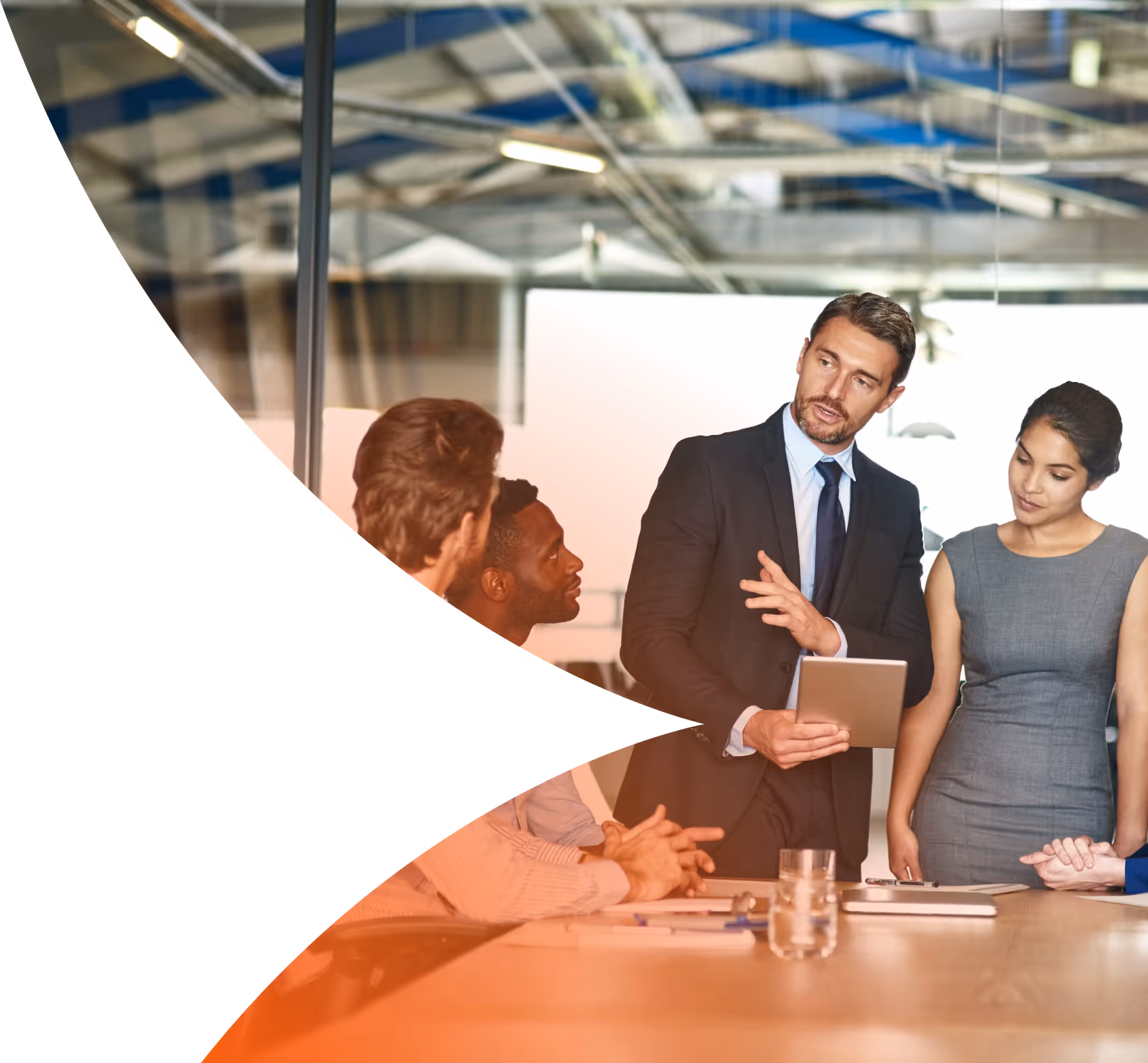 Businessman in a suit holding a tablet and speaking to colleagues around a conference table in a modern office.
