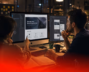 Two designers reviewing brand experience and website layouts on dual monitors in a dimly lit workspace.