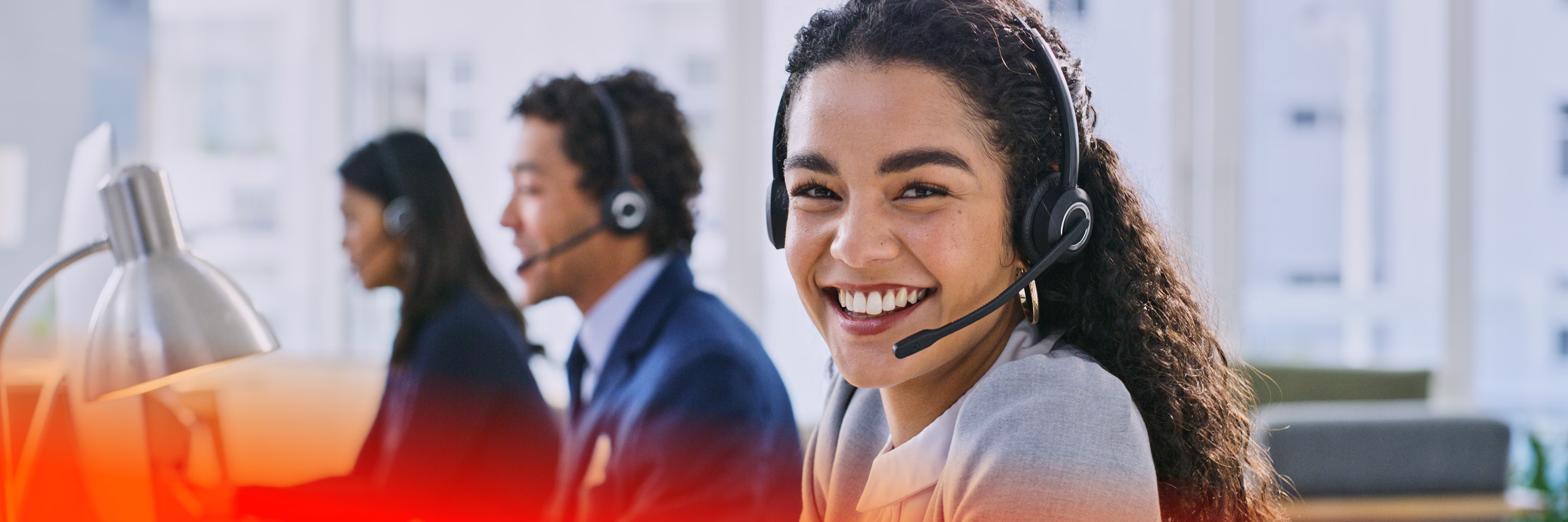 Smiling woman wearing headset at a call center with two colleagues working in background.