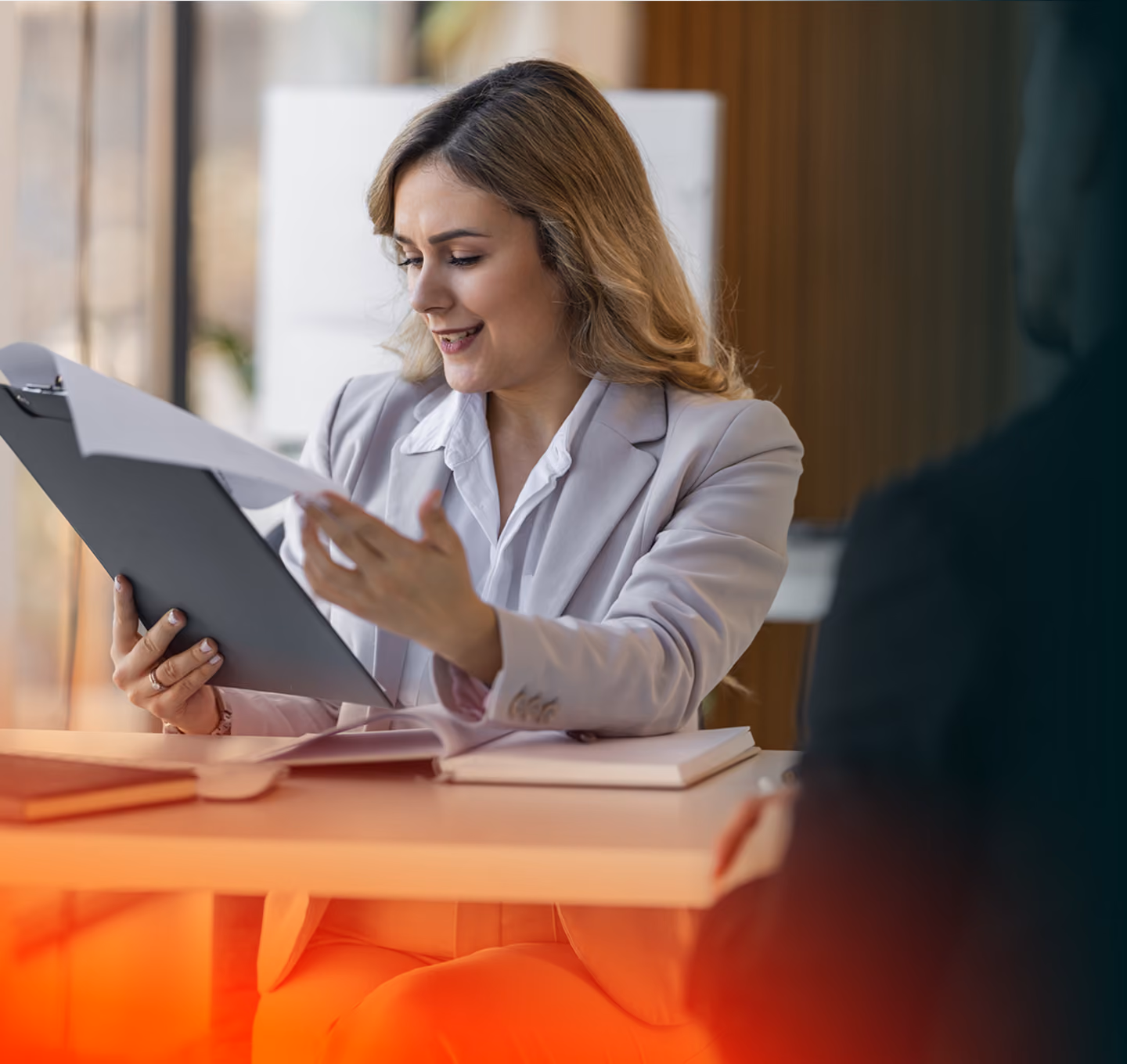 Smiling woman in a blazer holding a clipboard, reviewing documents at a desk in an office.