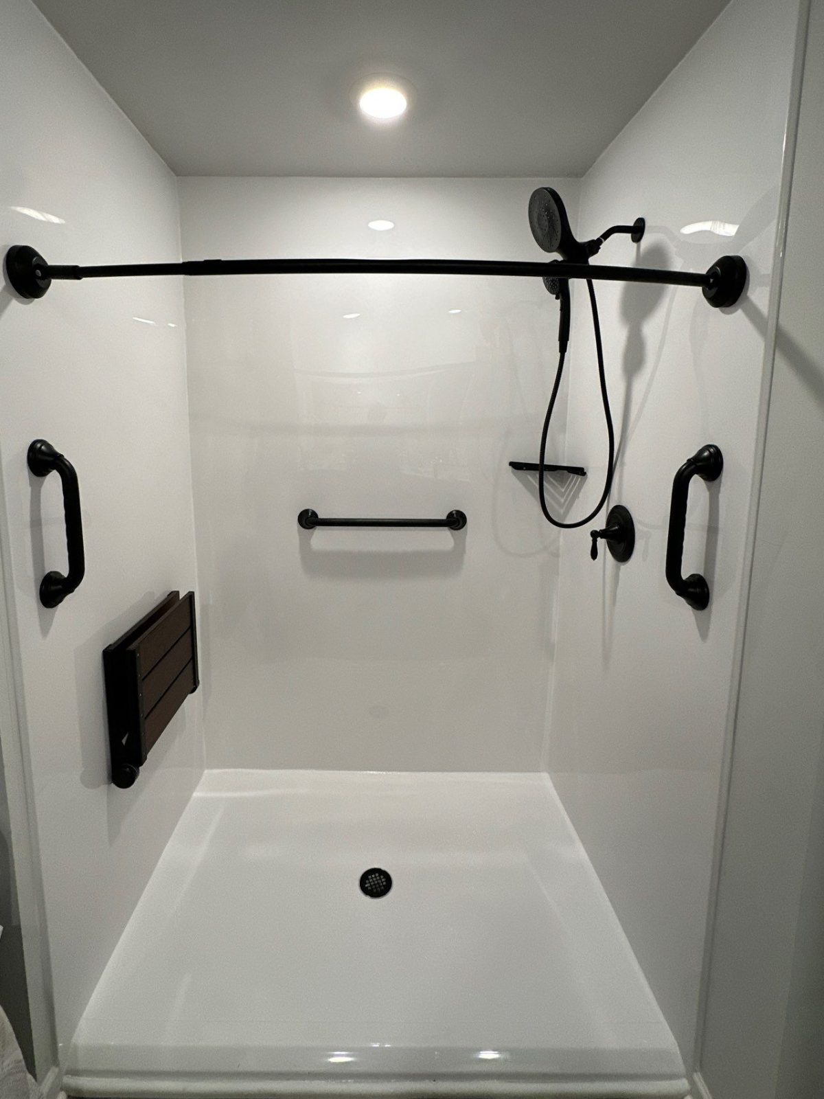 Same Baxter bathroom after shower replacement with a white zero-barrier shower, fold-up seat, matte black grab bars and a curved curtain rod