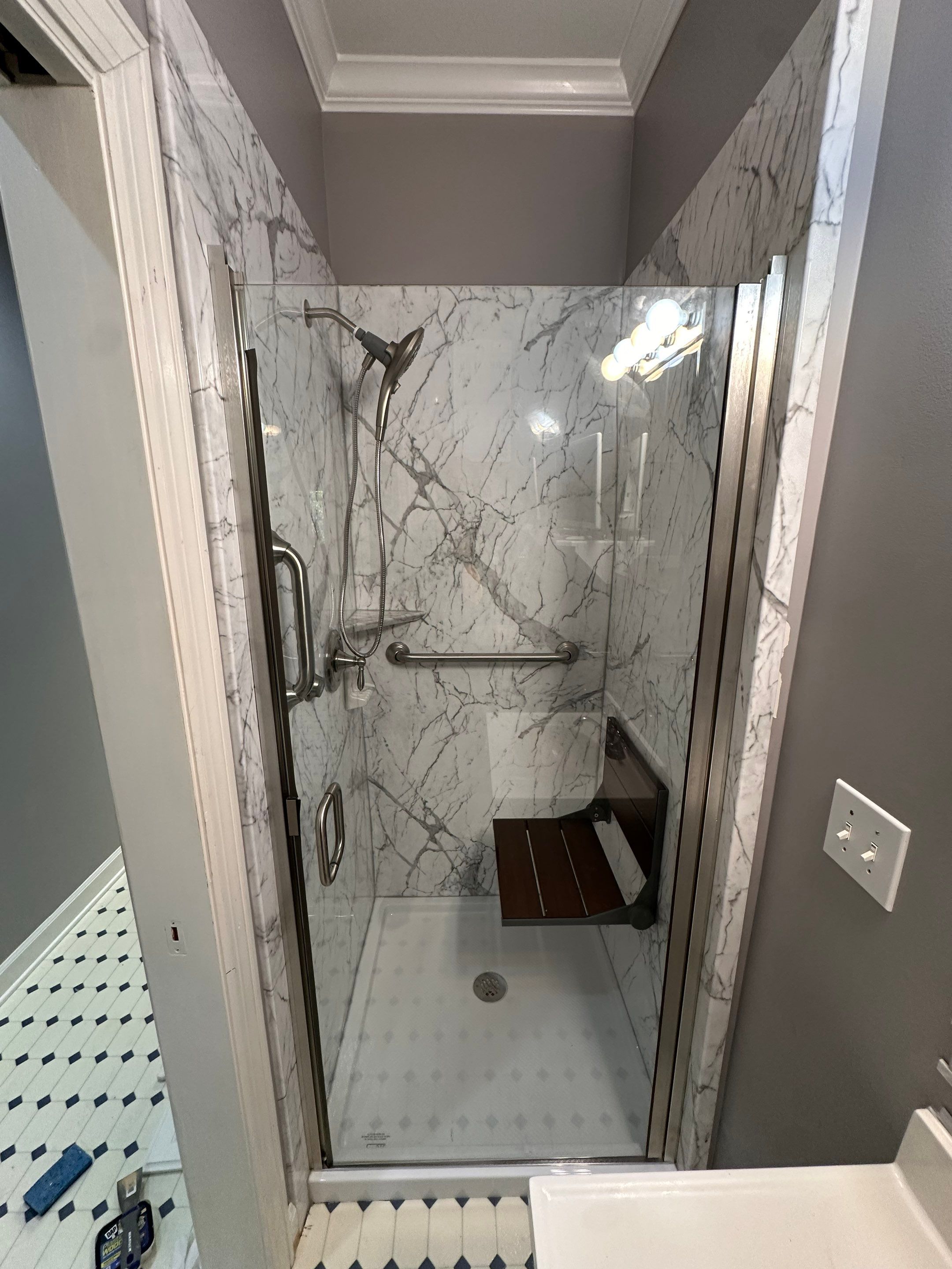 Same Nashville bathroom after shower replacement with gray-veined marble-look walls, a fold-up wood seat, grab bar and brushed nickel pivot glass door