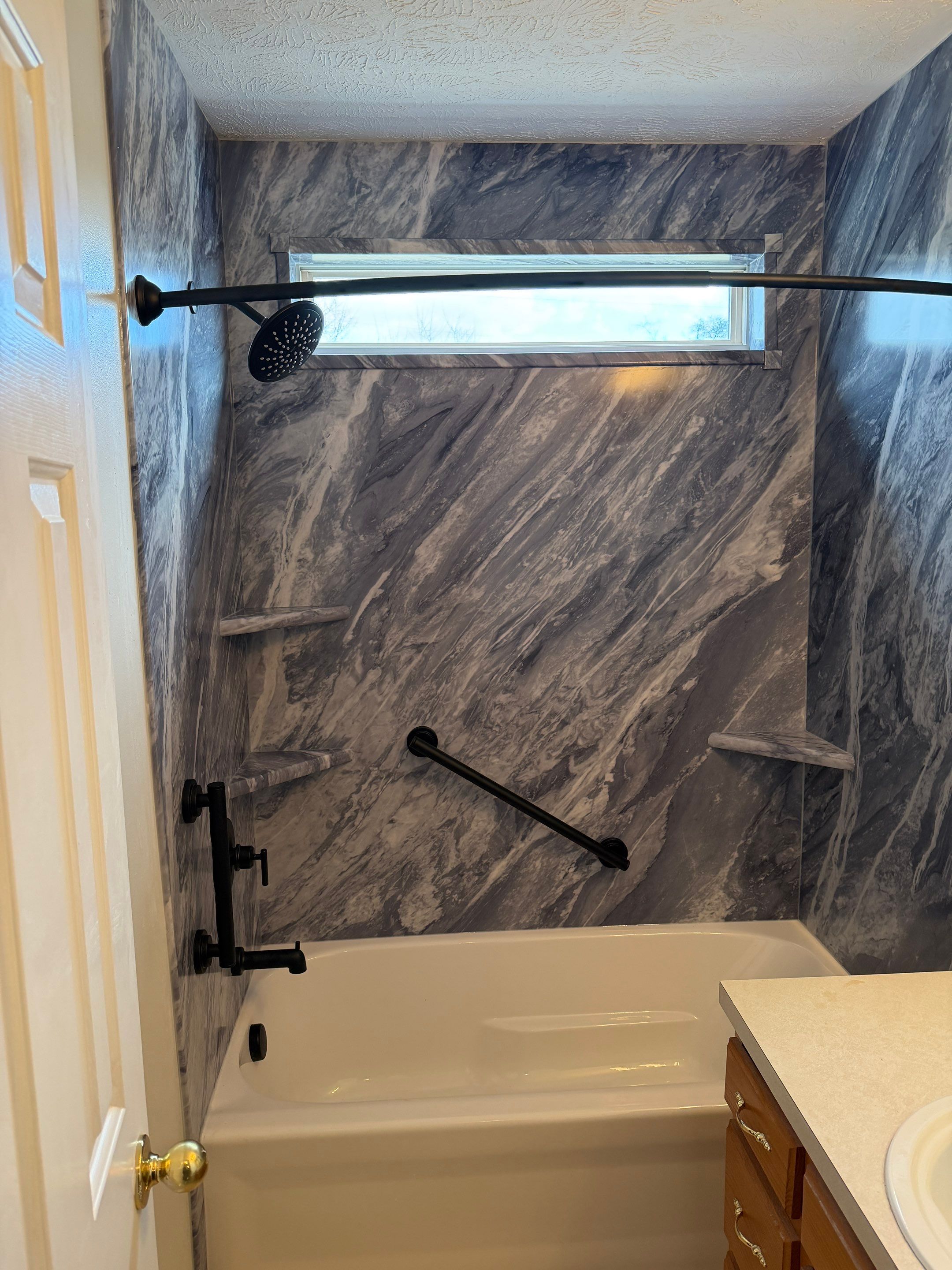 Same Antioch bathroom after tub replacement with a deep white bathtub, gray and white marble-look walls, a trimmed-in window, matte black fixtures, grab bar, corner shelves and a curved curtain rod