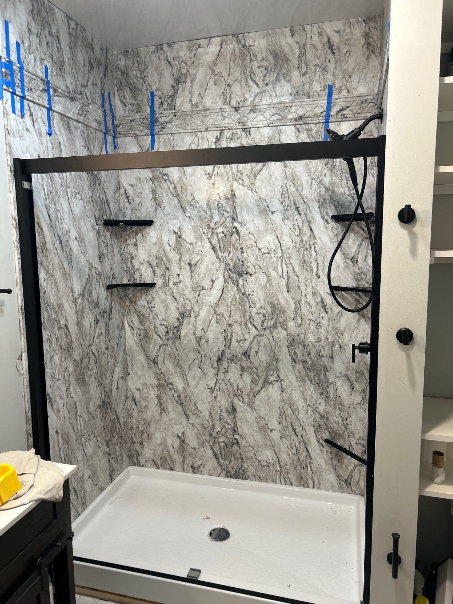 Same Bell Buckle bathroom after shower replacement with gray and white marble-look walls, a white base, matte black handheld shower, corner shelves and a matte black barn-style door frame