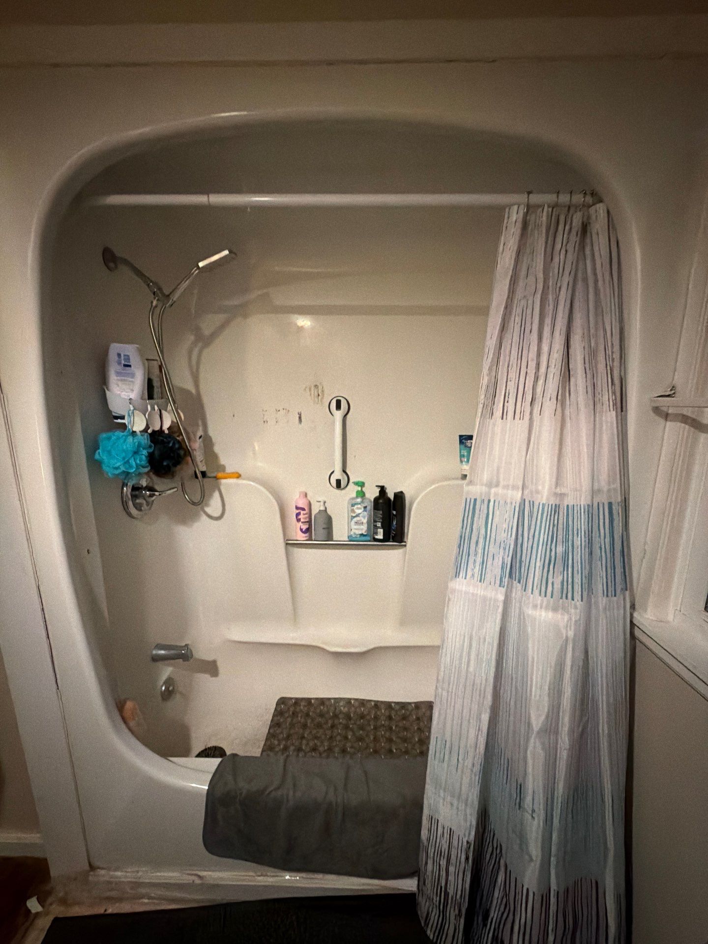 Worn white fiberglass tub and shower combo with a suction-cup grab bar, stained walls and a striped curtain before tub-to-shower conversion in Madison, TN