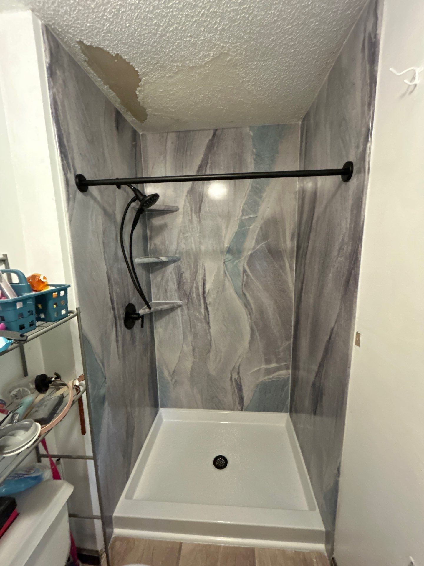 Same Clarkrange bathroom after shower replacement with gray, blue and violet stone-look walls, a white base, matte black handheld shower and valve, matching corner shelves and a matte black curtain rod