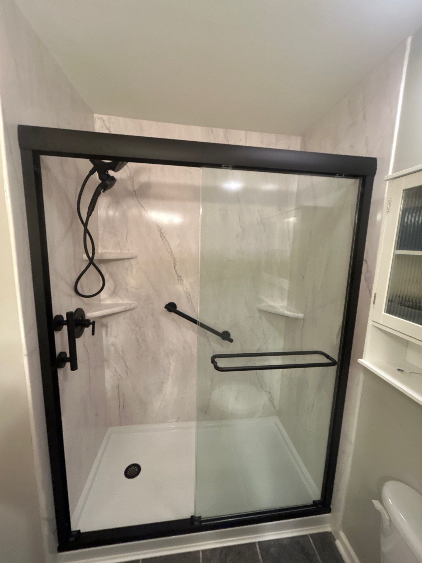 Same Spring Hill bathroom after tub-to-shower conversion with white marble-look walls with soft gray veining, a white base, matte black sliding glass door, handheld shower, grab bar and corner shelves