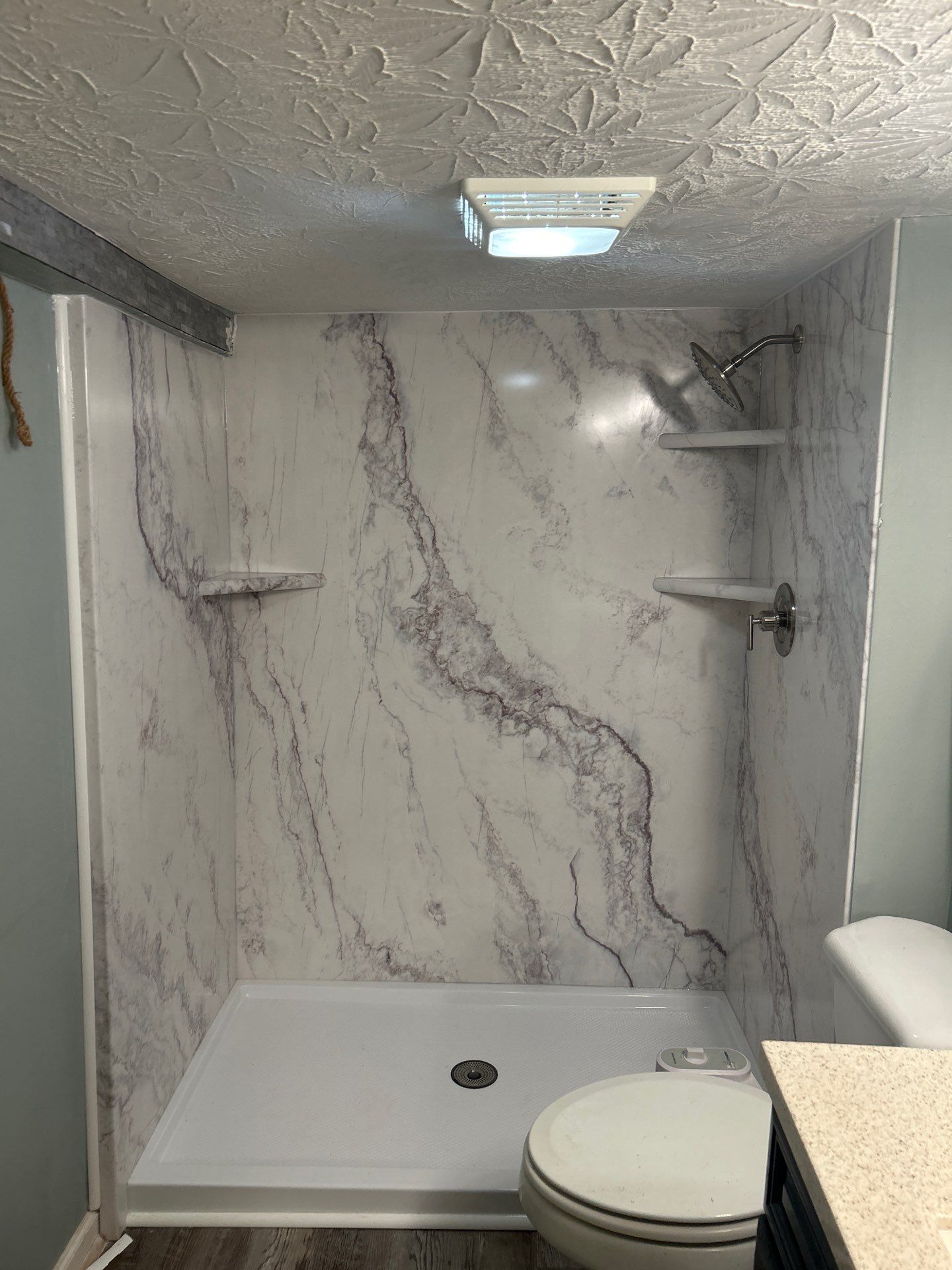 Same Westmoreland bathroom after shower replacement with white marble-look walls with soft gray and plum veining, a white shower base with center drain, three matching corner shelves and brushed nickel shower head and valve