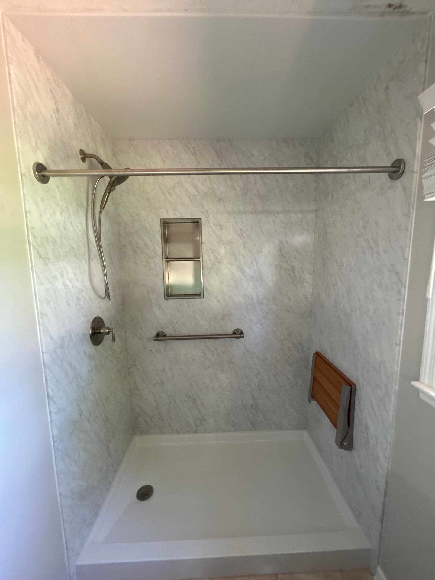 Same Winchester bathroom after tub-to-shower conversion with white and gray marble-look walls, a white low-threshold shower base, built-in niche shelves, teak fold-down seat, grab bar, handheld shower and brushed nickel curtain rod