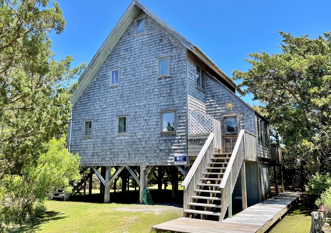 Cedar-shingle beach house elevated on stilts with exterior stairs leading to the main entrance