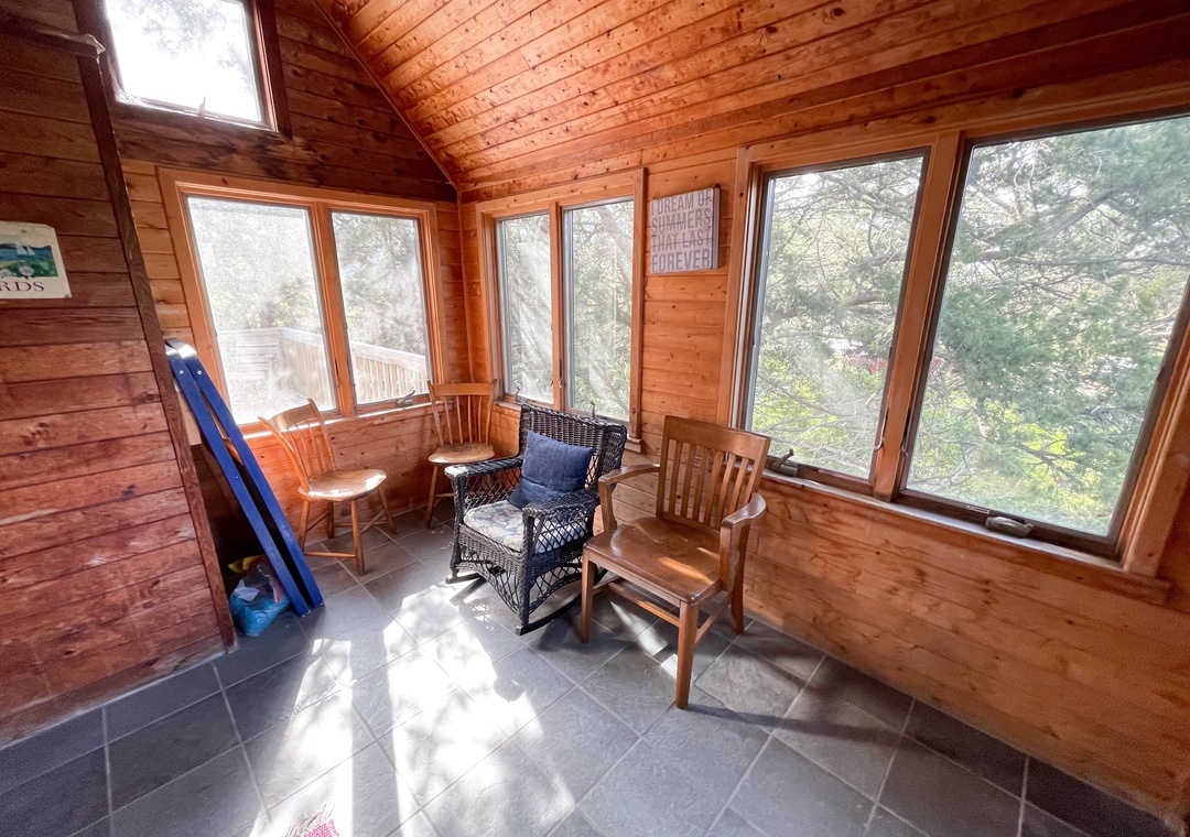 Enclosed sunroom with wood walls and ceiling, tile floor, and a mix of wooden and wicker chairs facing multiple windows