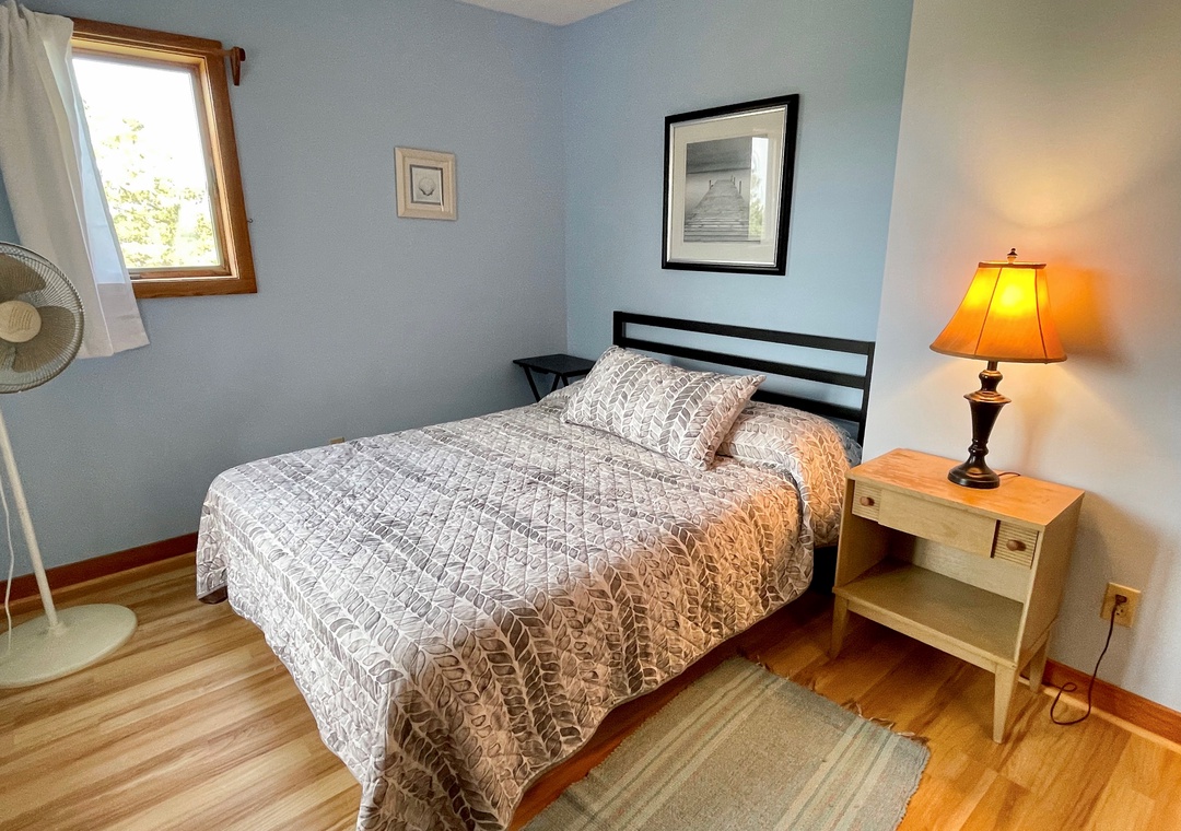Bedroom with a full-size bed, gray patterned quilt, light blue walls, small nightstand, and window with white curtains