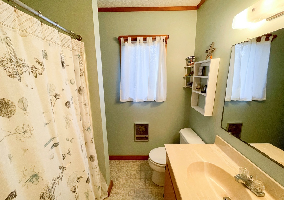 Small bathroom with light green walls, a floral shower curtain, single vanity, and window with white curtains