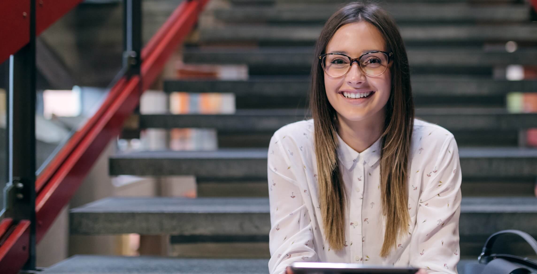Smiling young woman with glasses sitting on stairs holding a tablet.
