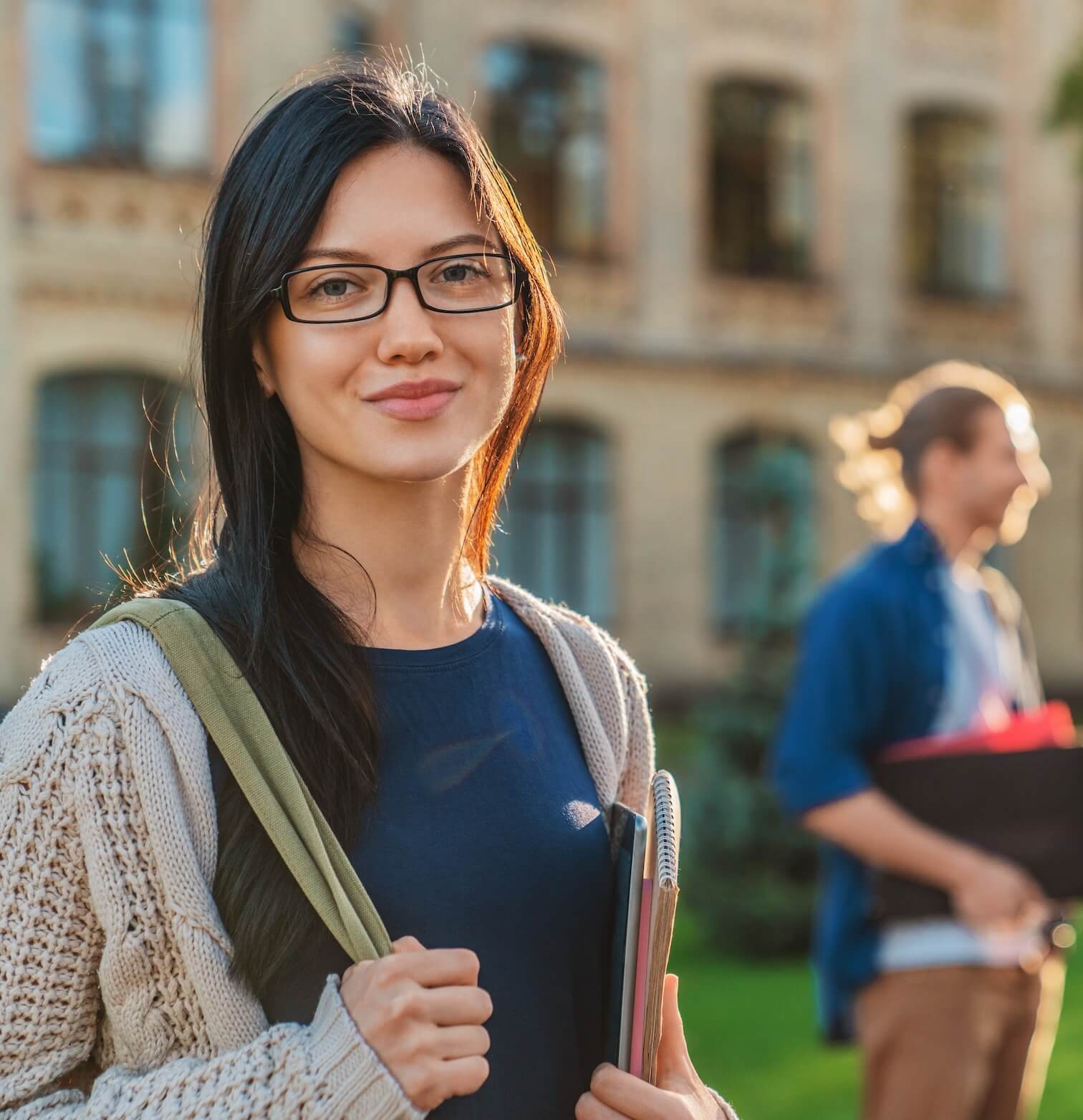 Smiling young woman with glasses holding notebooks and a backpack standing outside a building, with a blurred man in the background.