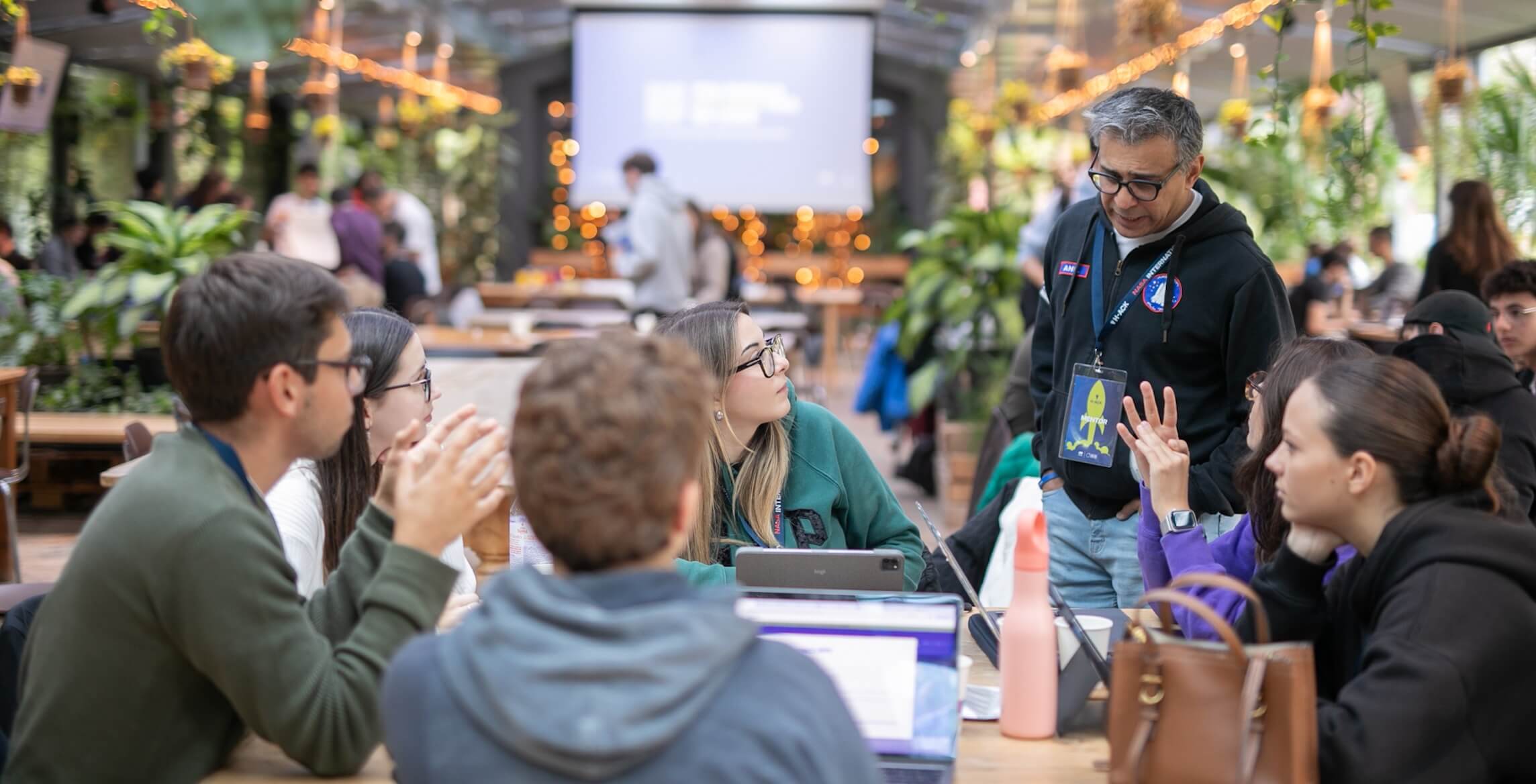 A group of students sitting around a table with laptops and tablets, engaged in discussion with a standing mentor in a lively, plant-filled indoor space.