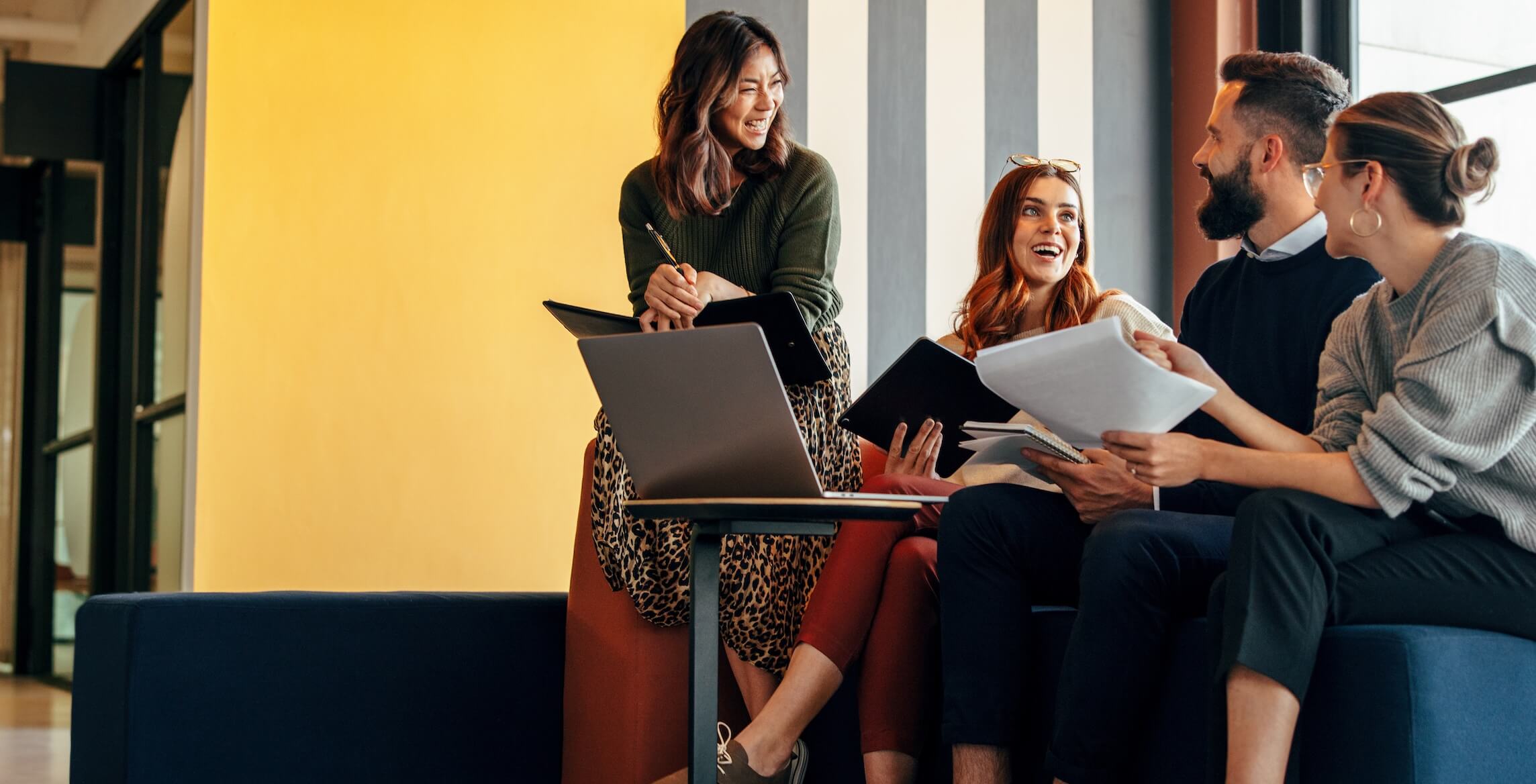 Four colleagues smiling and discussing documents while sitting on a blue couch with a laptop in a modern office.