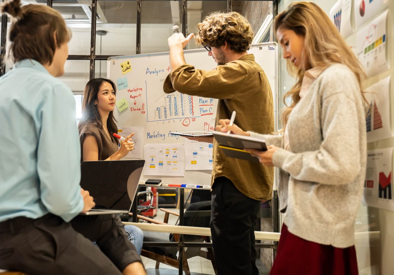 Four young professionals collaborating in an office with charts and notes on a whiteboard and walls, discussing a marketing report.