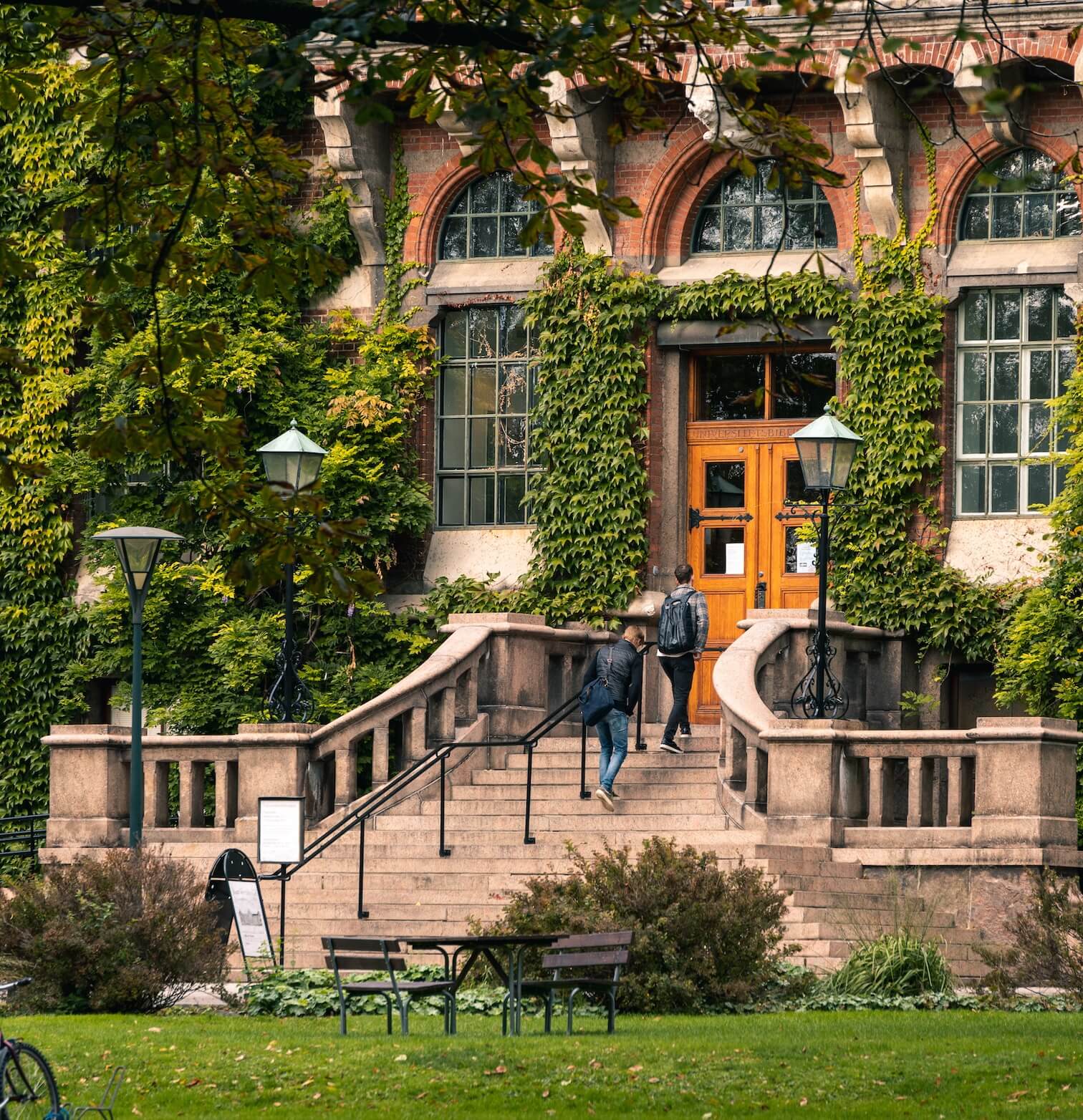 Two people walking up stone steps to a wooden door of a brick building covered in green ivy.