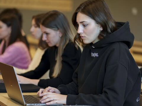 Young woman in a black hoodie focused on typing on a laptop with two other women working on laptops in the background.