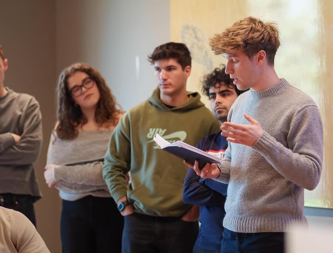 Young man in a gray sweater reading from a notebook while others listen attentively in a casual indoor setting.