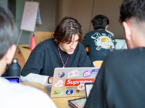 A young man with wavy brown hair focused on his laptop covered with stickers, sitting at a table with two other people working nearby.