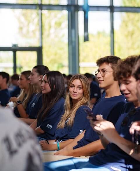 A group of young people sitting indoors in rows, wearing matching navy blue shirts, with one woman looking directly at the camera.