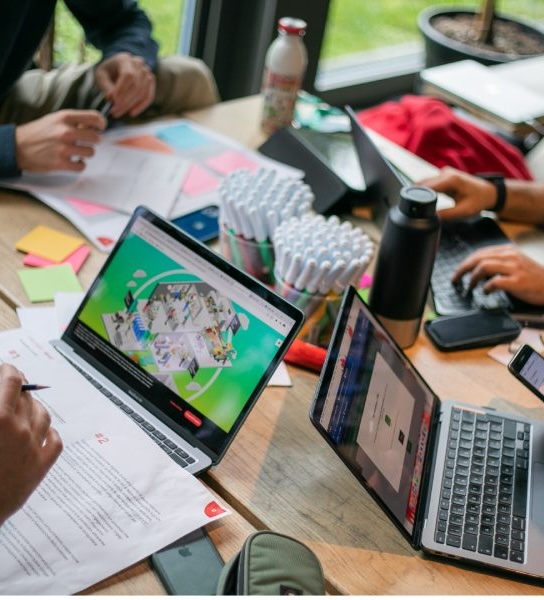 People working collaboratively at a wooden table with laptops, documents, markers, and smartphones.