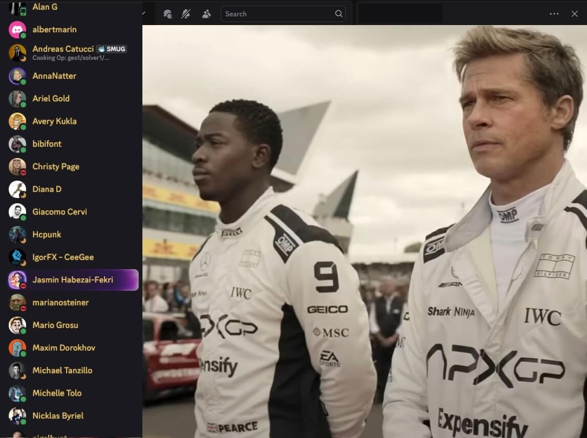 Two race car drivers in white racing suits standing side by side at a race track with a racing car and crowd in the background.