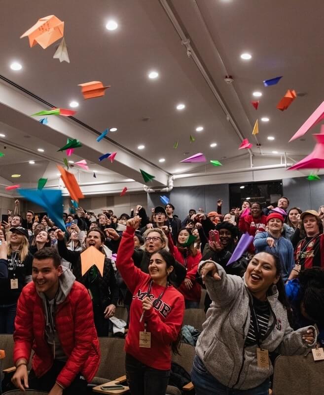 A diverse group of enthusiastic people indoors throwing colorful paper airplanes into the air.
