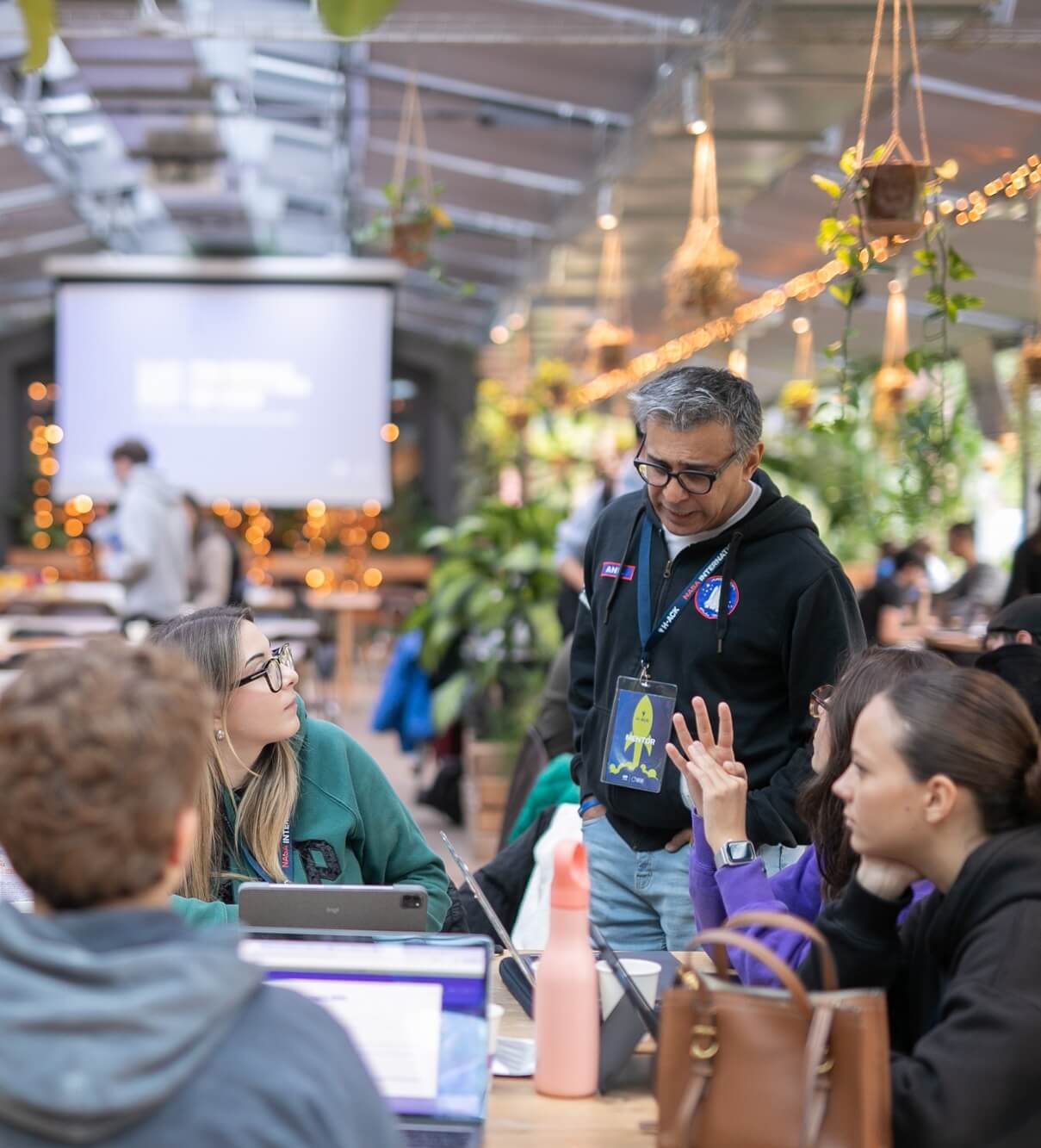 A man wearing glasses and a mentor badge talks to three seated young adults with laptops in a brightly lit indoor space decorated with hanging plants.