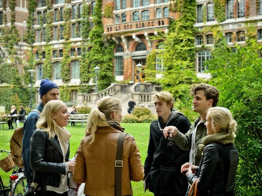 A group of six young adults talking outdoors in front of a large ivy-covered brick building on a grassy area.
