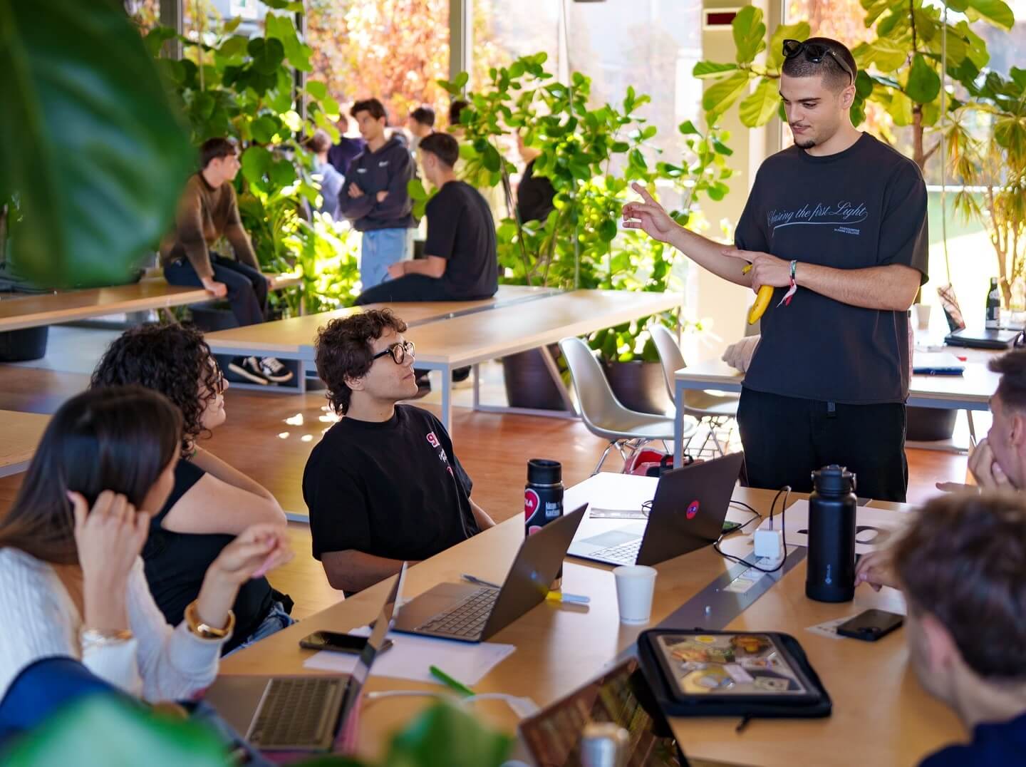 Group of young adults engaged in a discussion around tables with laptops in a bright, plant-filled room.