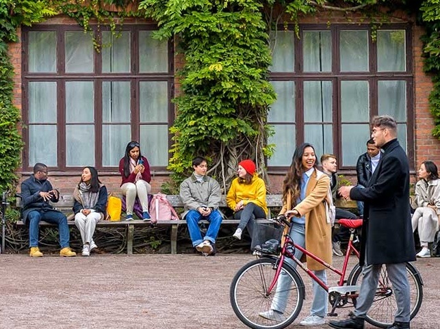 Group of diverse young adults sitting and chatting on a wooden bench under windows with ivy, while a woman with a bicycle and a man in a coat talk nearby.