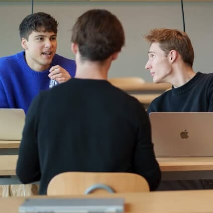Three young men sitting at a table, two facing each other with laptops, engaged in conversation.