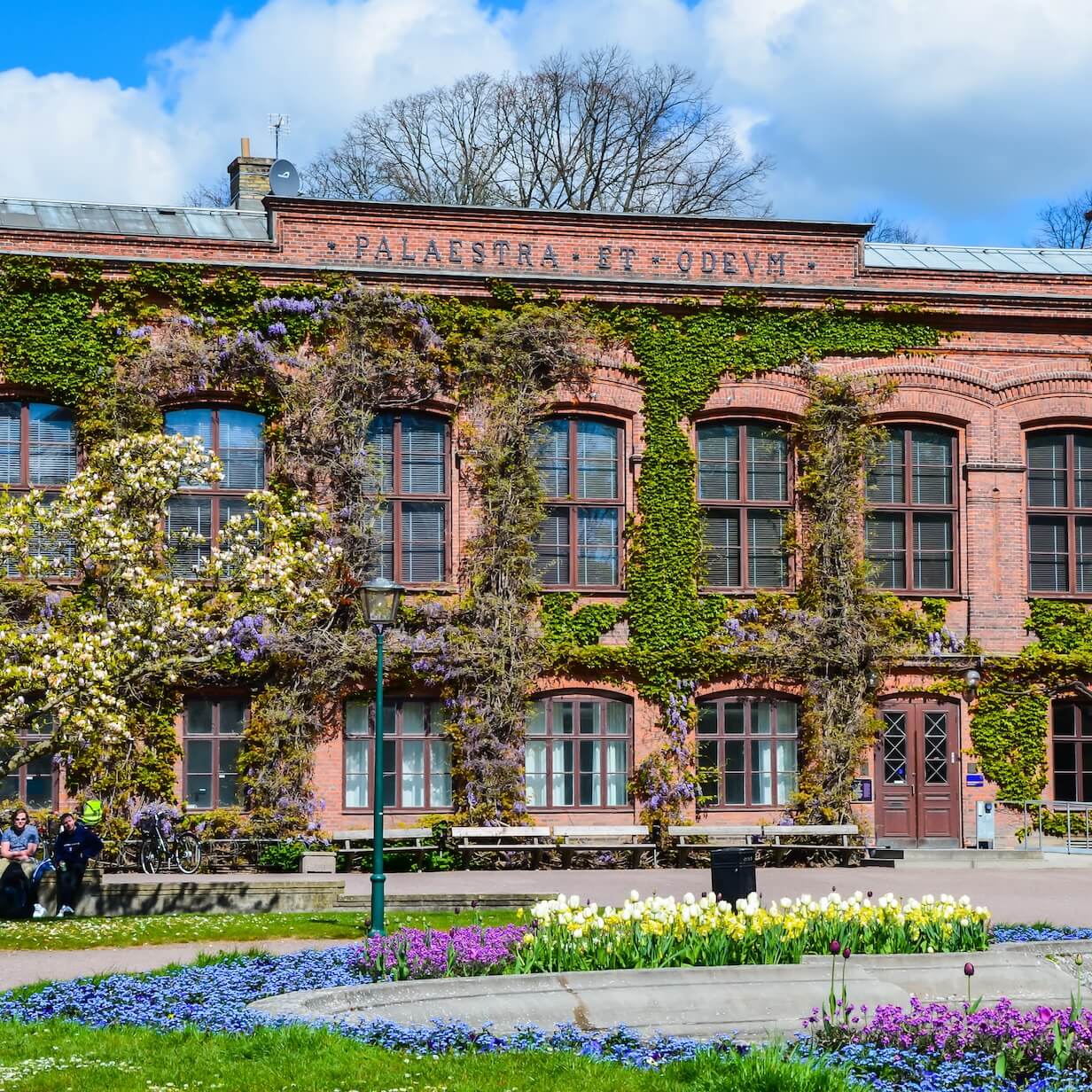 Brick building covered in ivy with large windows and a Latin inscription, in front of a garden bed of colorful flowers and a lamppost.