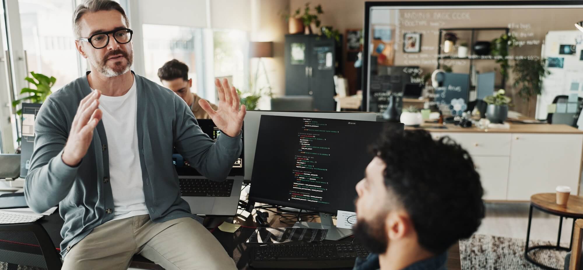 A man with glasses explaining code on a computer screen to a seated colleague in a modern office.