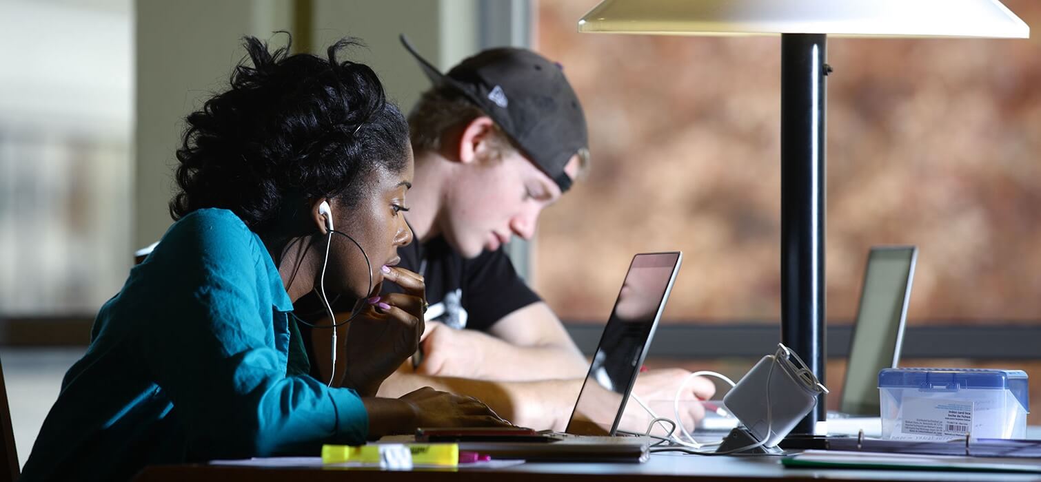 Two students focused on laptops studying at a table with a desk lamp.