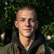 Young man with short hair wearing a green jacket outdoors with leafy background.