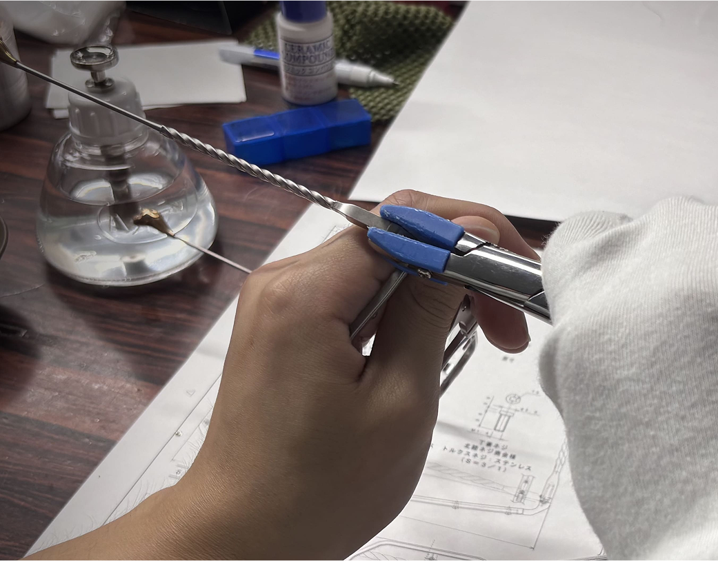A white-gloved artisan using a precision hand-tool to meticulously assemble the signature twisted arm of a DRK frame, with technical schematics visible.
