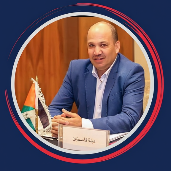 A man in a blue blazer seated at a desk with a Palestinian flag and a nameplate in Arabic.