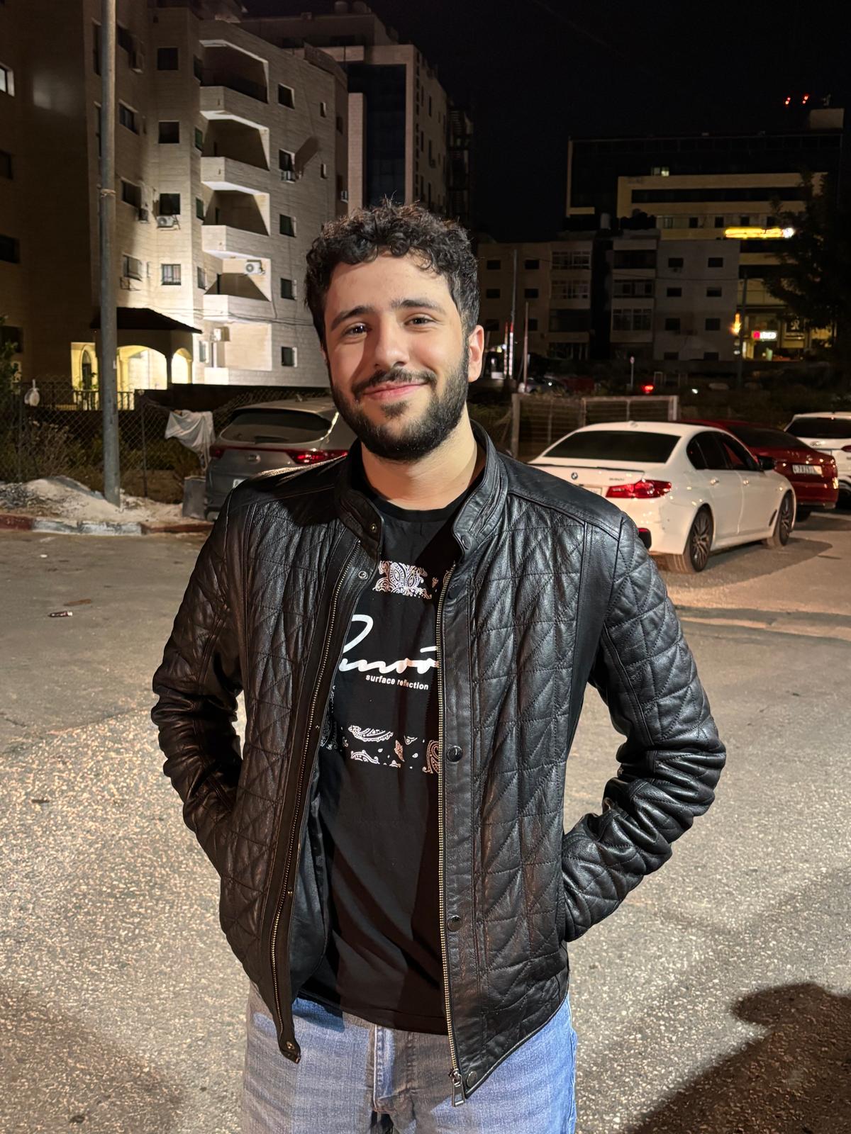 Smiling man with curly hair and beard wearing a black quilted leather jacket and jeans standing outdoors at night.