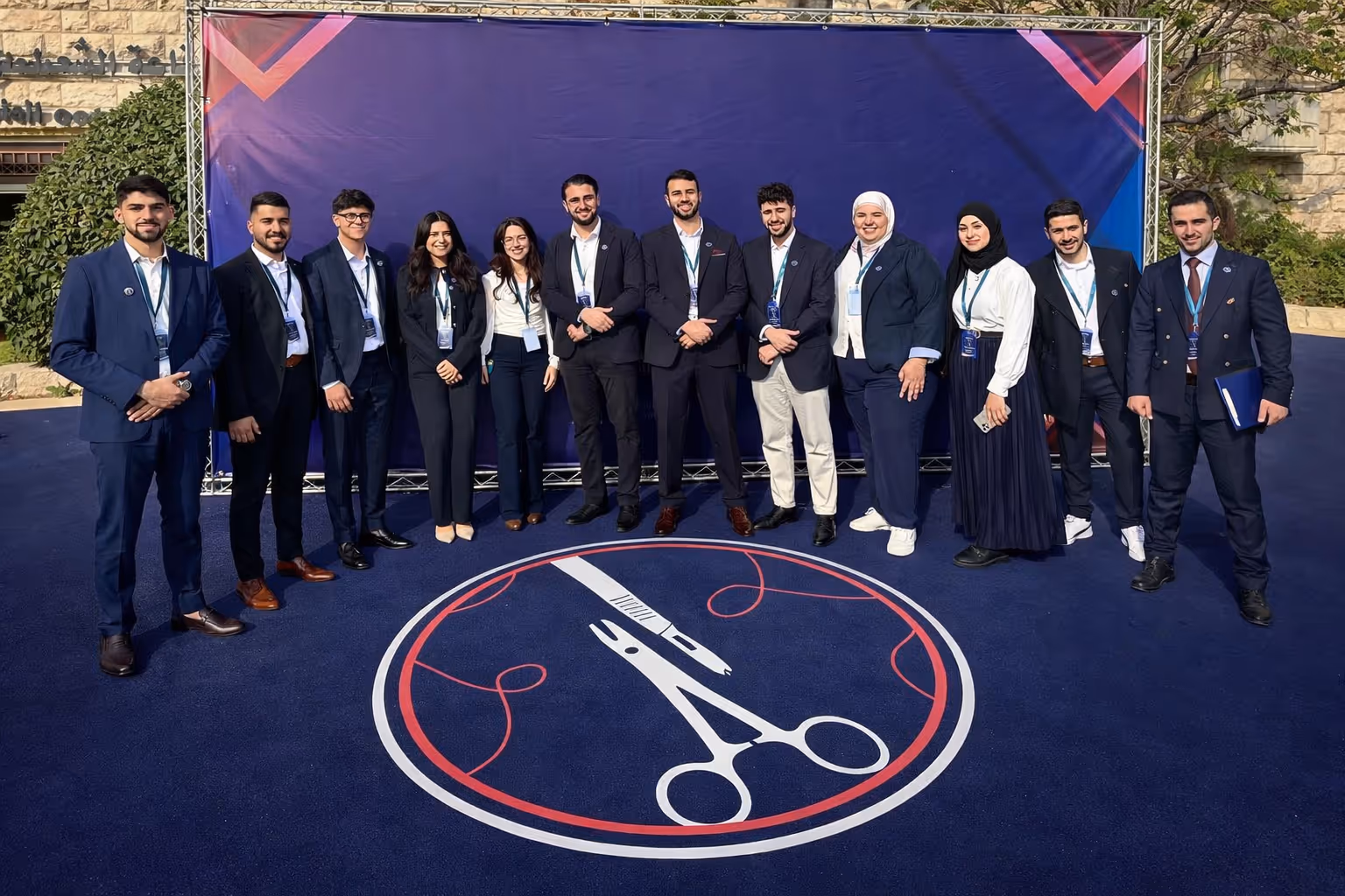 Group of eleven young adults dressed in formal attire standing on a blue carpet with a surgical scissors and thread symbol, posing in front of a large purple backdrop.